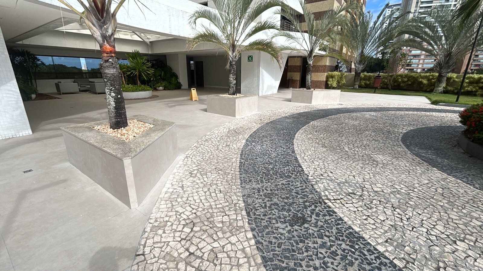 Transição entre piso claro em placas e mosaico de pedras na área externa do Condomínio Lumno, com jardineiras e palmeiras.