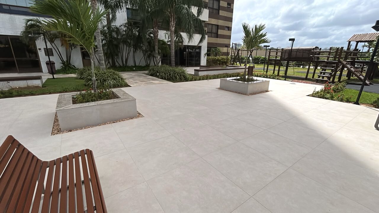 Praça externa do Condomínio Lumno com piso claro em placas, jardineiras e playground ao fundo.